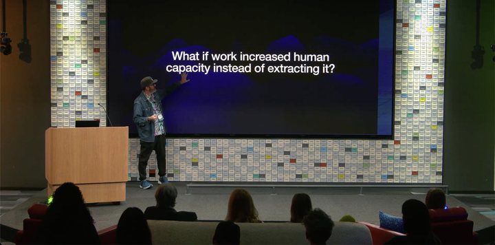 Mike McHargue on stage at Google, gesturing toward a large screen that reads "What if work increased human capacity instead of extracting it?" while a seated audience watches.